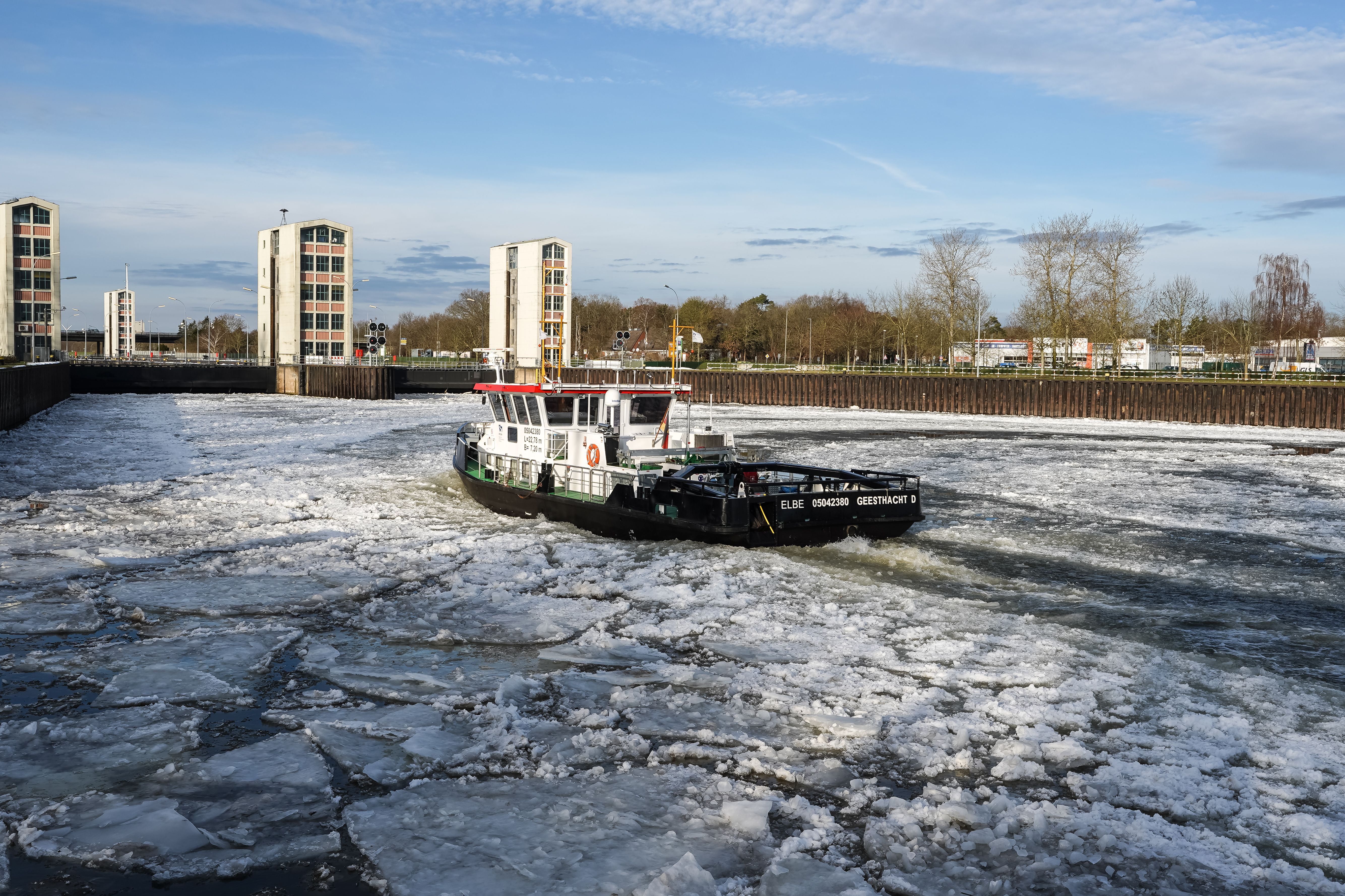 Ice-covered river with a small boat in the center and buildings along the shore under a blue sky