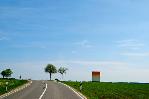 一条宁静的乡村道路，树木稀疏，蓝天下矗立着一栋建筑