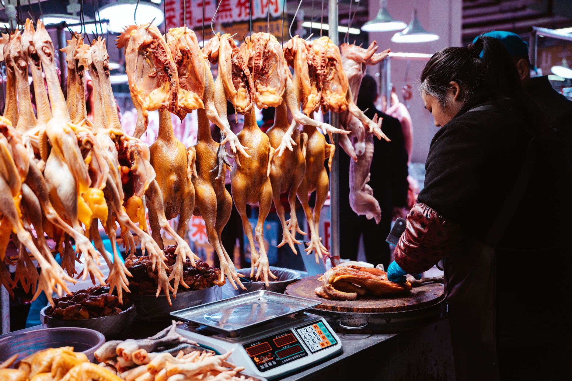 A market vendor prepares poultry at a traditional indoor Asian marketplace with hanging chickens.