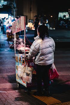 A street vendor with food cart operating at night in a busy city setting. Captured from behind, highlighting urban lifestyle.