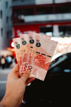 Close-up of a hand holding Hong Kong dollar banknotes against a vibrant city background at night.