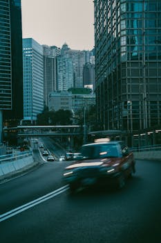 Blurred red car on a bustling city street surrounded by skyscrapers at dusk.