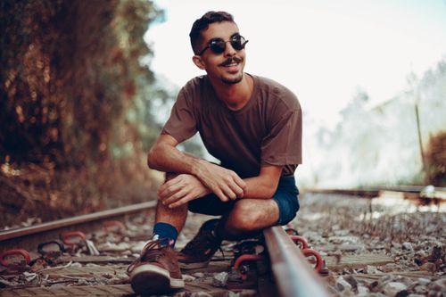 Free Fashionable man in sunglasses sitting on railway tracks, smiling on a sunny day. Stock Photo