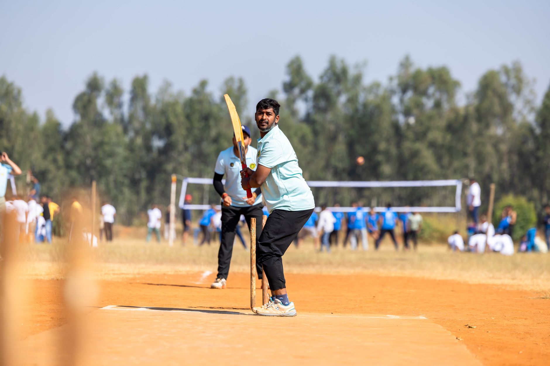 A cricketer prepares to bat during an outdoor game on a sunny day, with spectators in the background.