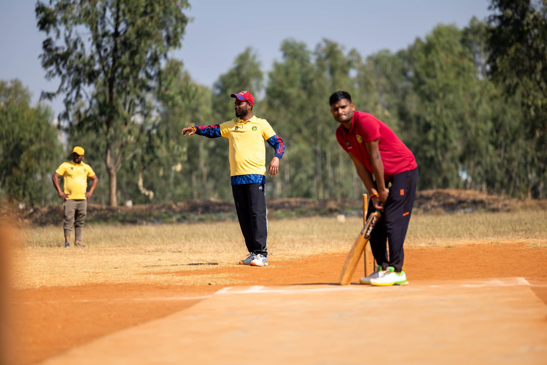Cricket players in action during an amateur match on a sunny day outdoors.