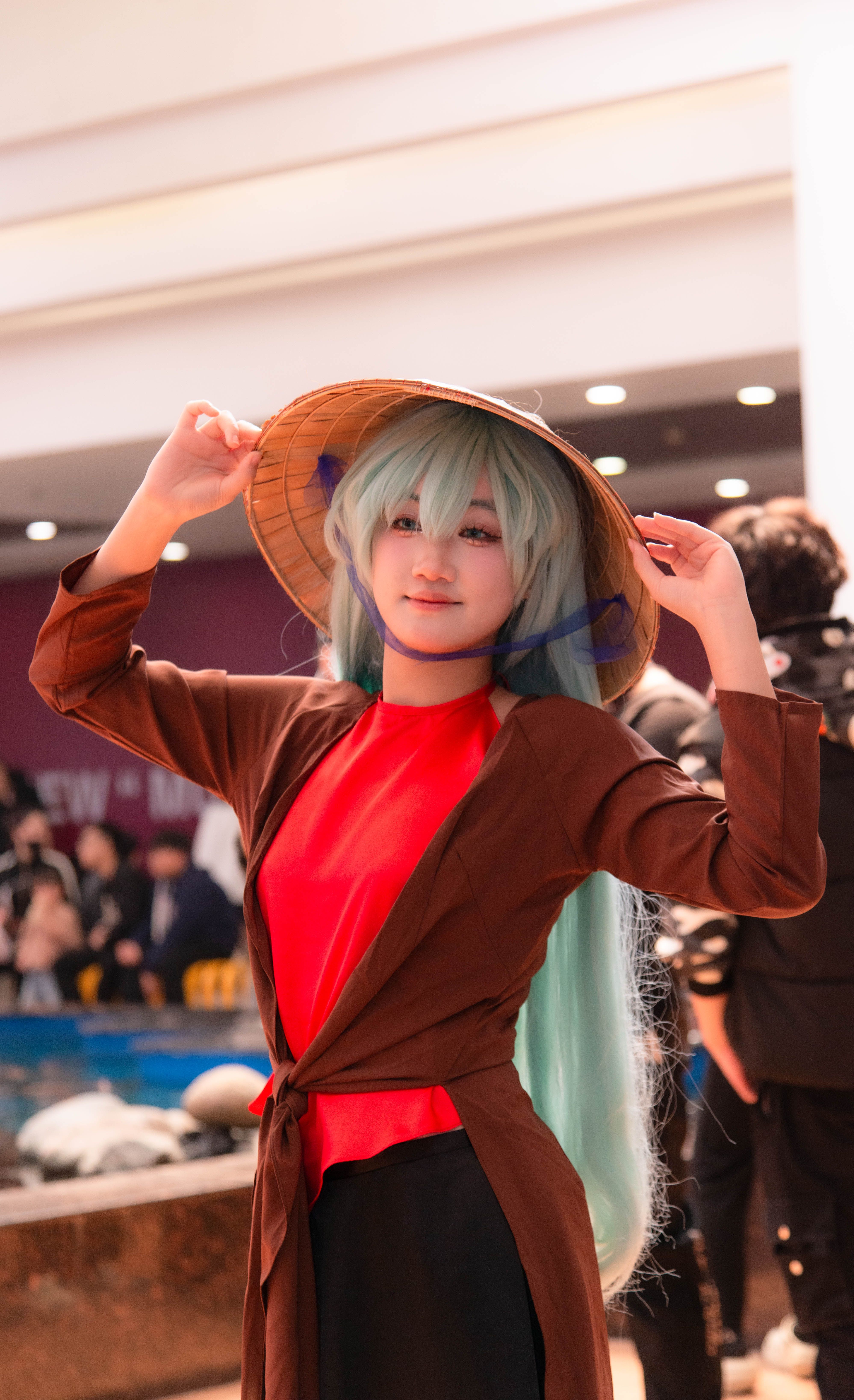 A cosplay enthusiast wearing traditional attire and a conical hat poses indoors at a lively event.