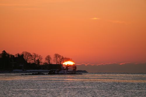 Free Beautiful winter sunset casting warm hues over a calm ocean with silhouetted trees. Stock Photo