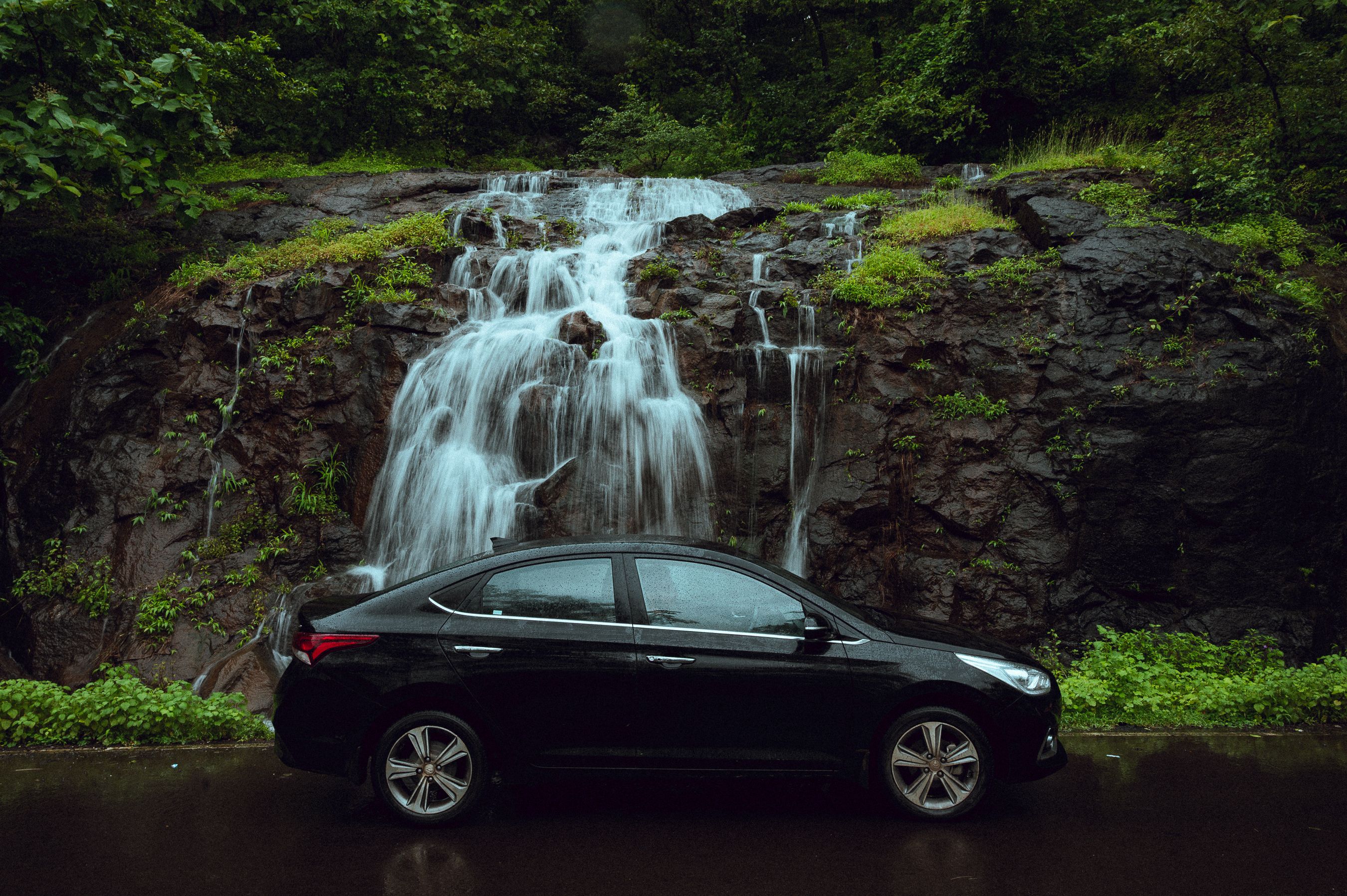 Black car parked in front of a serene waterfall surrounded by greenery in Mumbai, India.