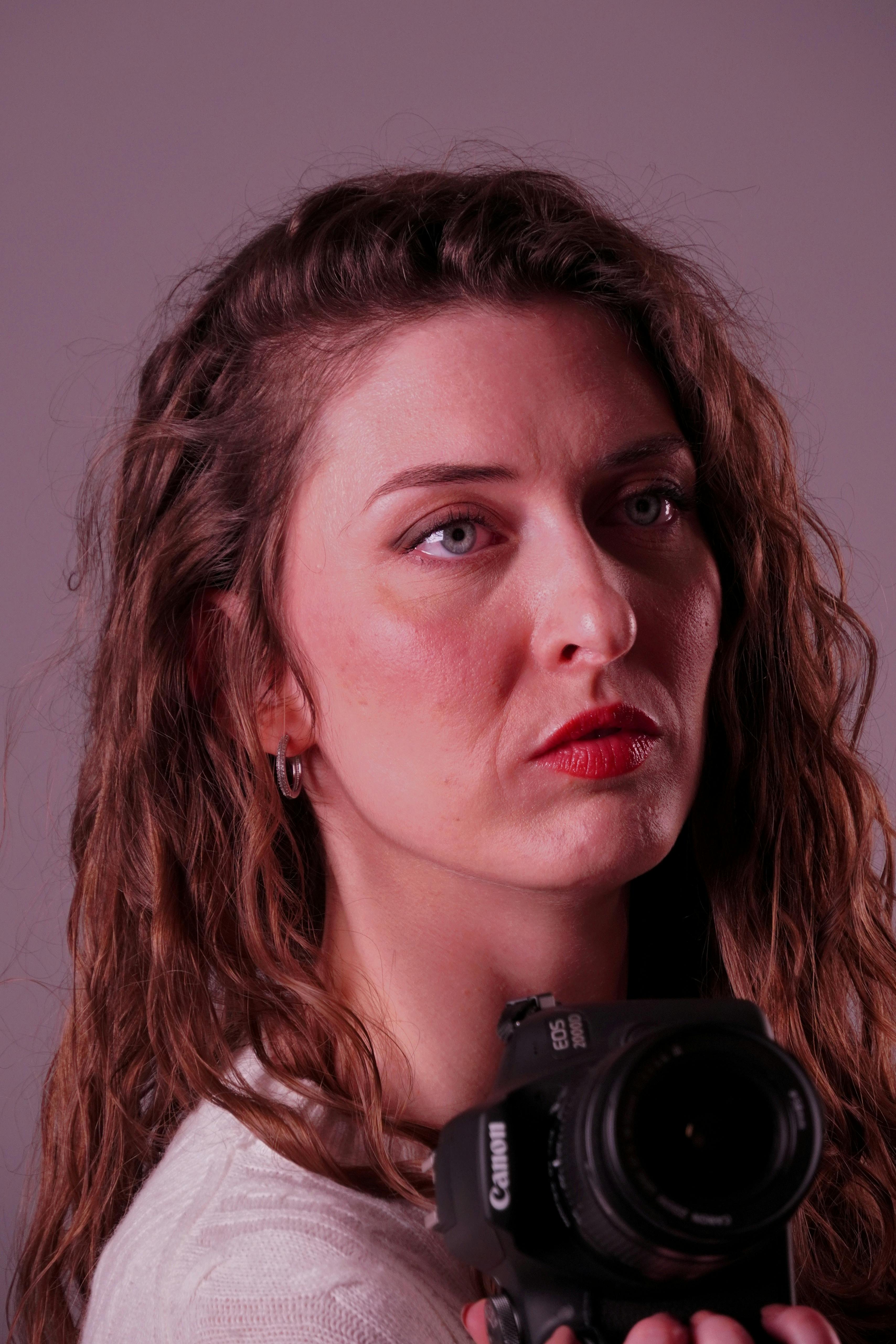 grátis Retrato em close-up de uma mulher com cabelos cacheados segurando uma câmera DSLR, em um fundo neutro. Foto profissional