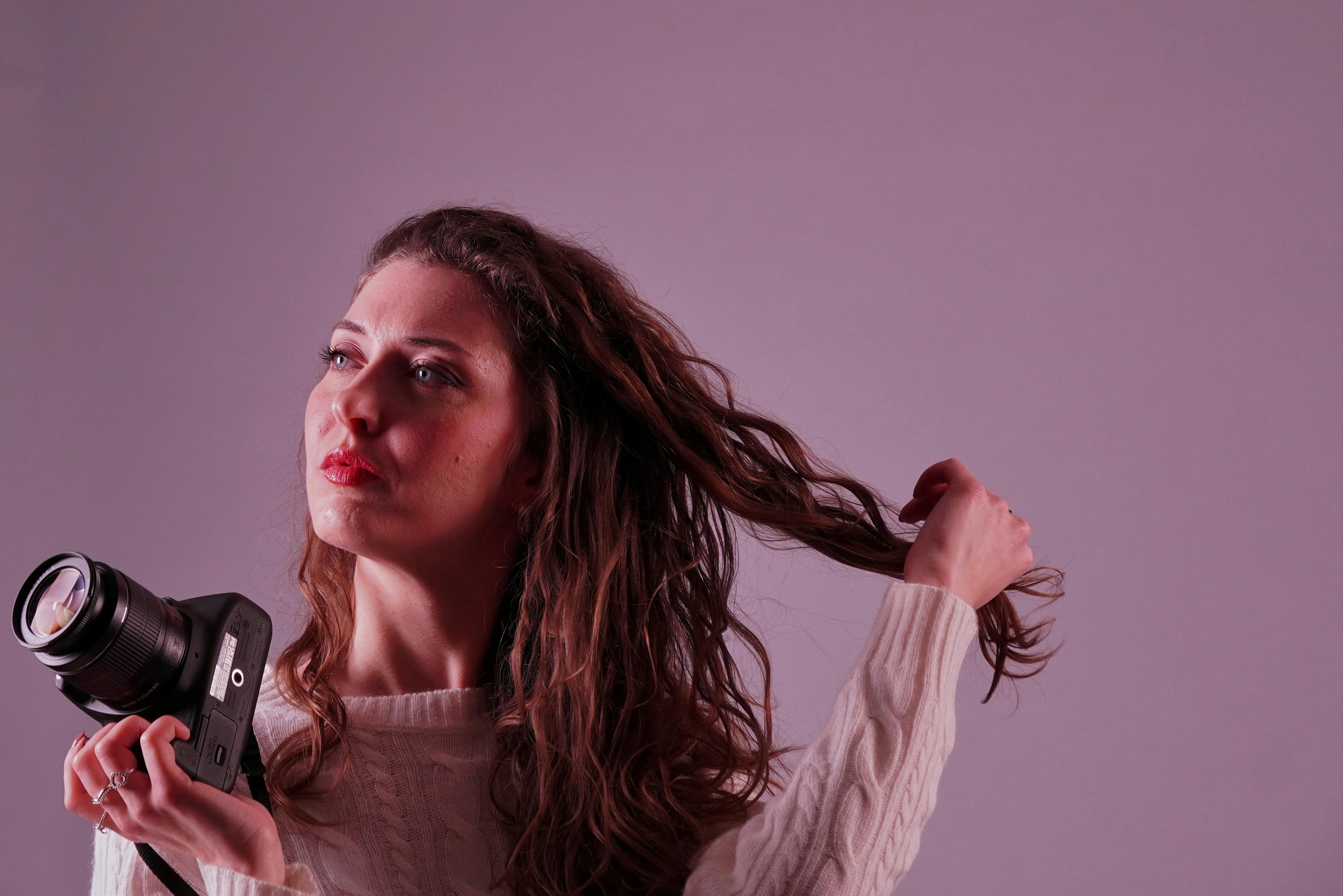 無料 白いセーターを着てカメラを持った女性を、暖かいスタジオの照明の下で撮影した写真。 写真素材