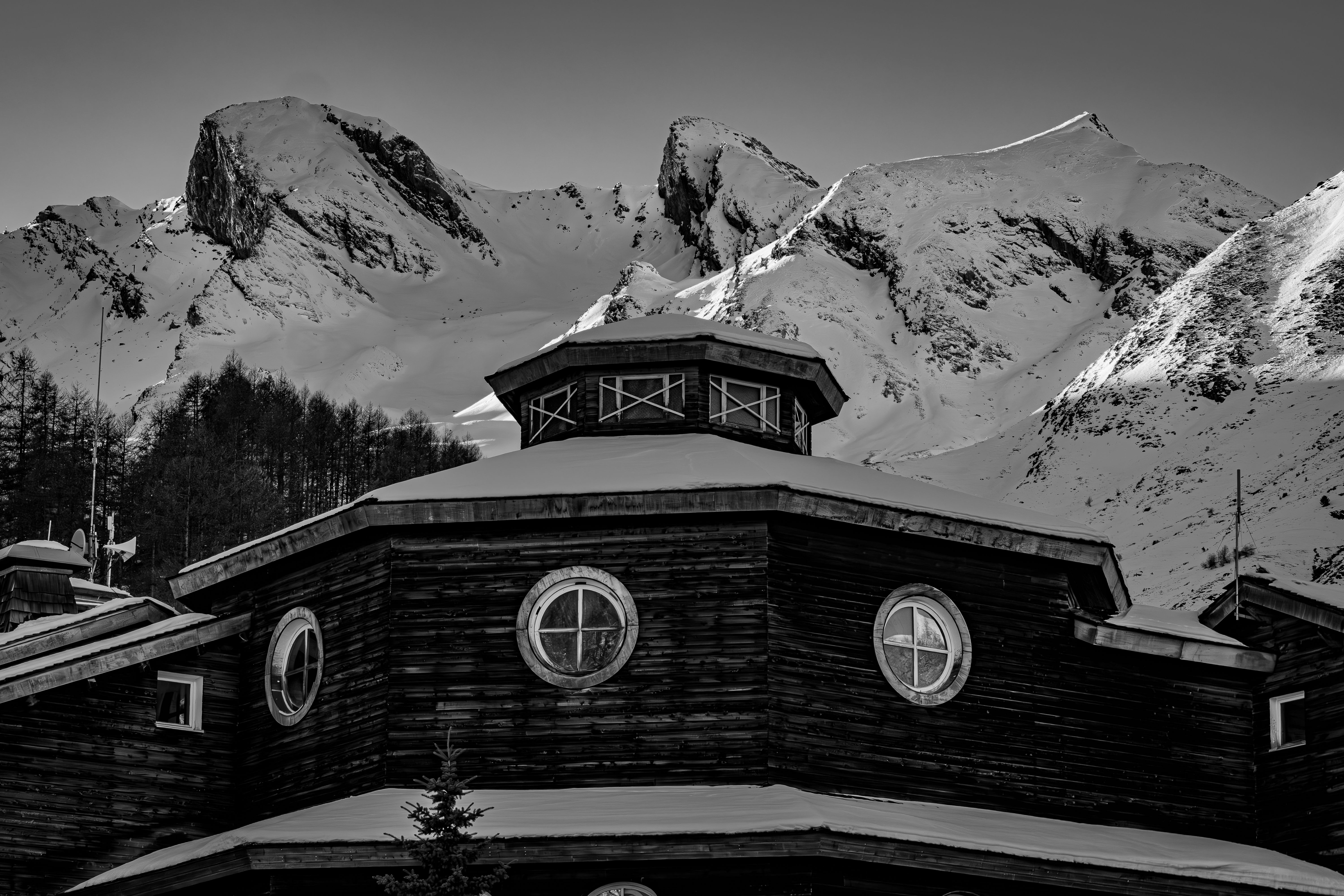 Une cabane rustique se détachant sur le fond de majestueuses montagnes enneigées, dans un paysage classique en noir et blanc.