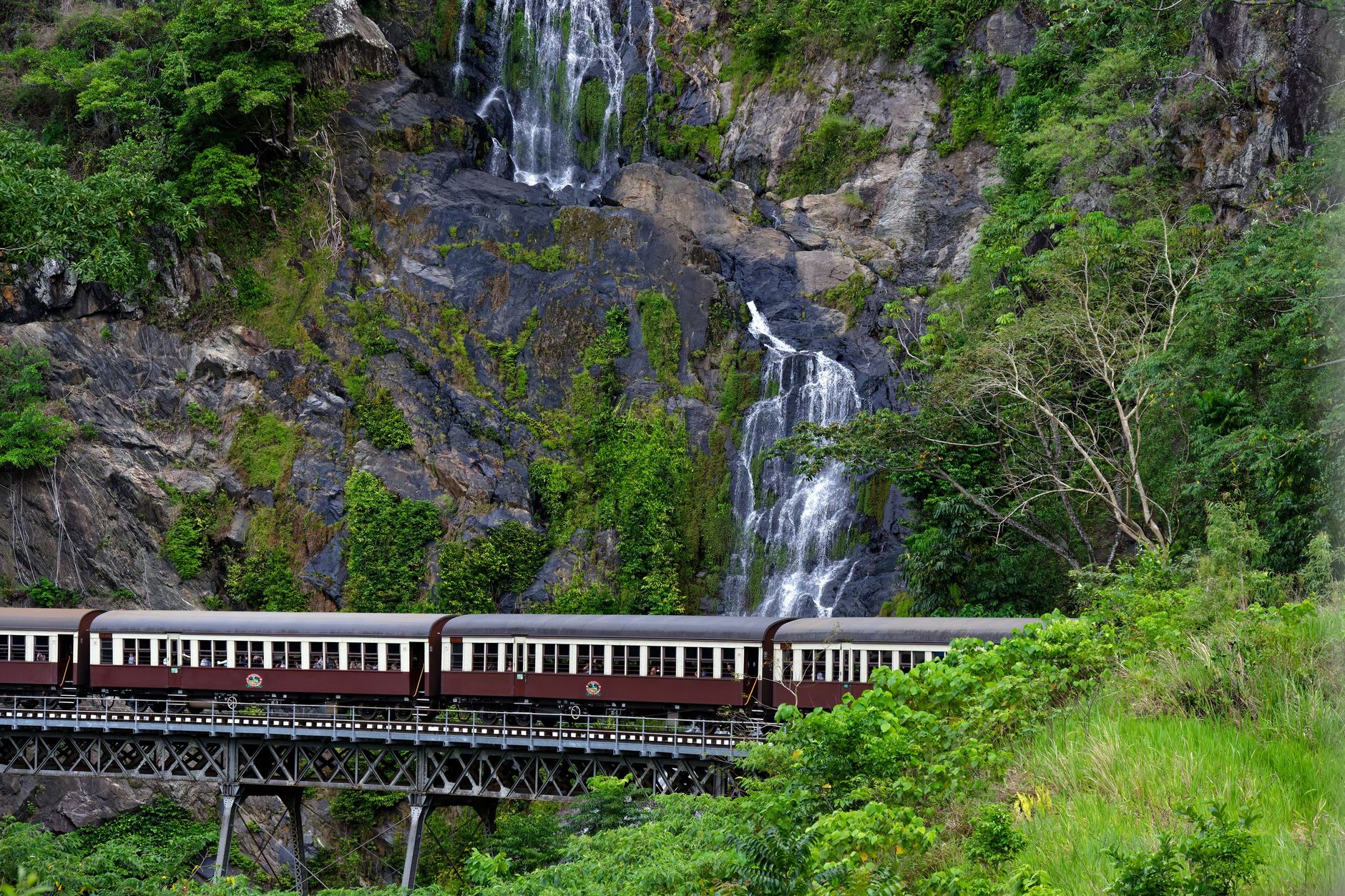 The maroon carriages of the Kuranda Scenic Railway crossing a trestle bridge below a waterfall in the Queensland rainforest