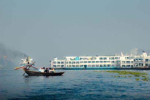 Free A traditional boat navigates the Buriganga River in Dhaka with a large passenger ferry in the backdrop. Stock Photo