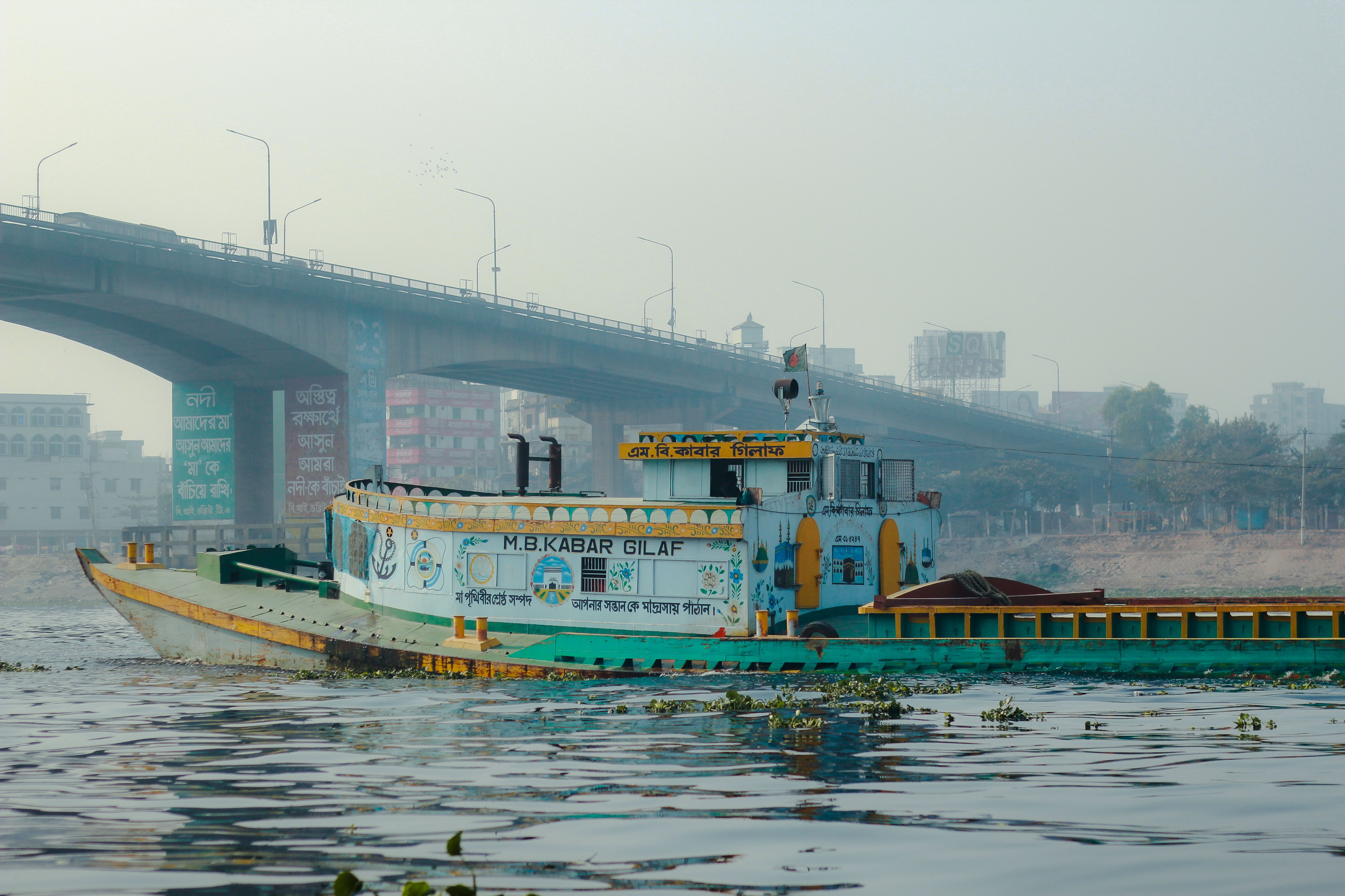 Vibrant boat sails along the Buriganga River, Dhaka, with cityscape backdrop.