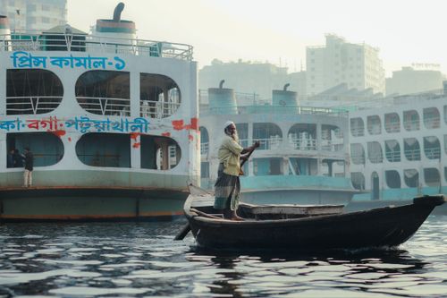 Free A man navigates his wooden boat through Dhaka's bustling Sadarghat, surrounded by large ferries. Stock Photo