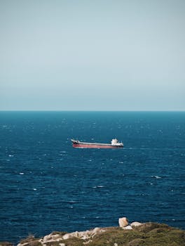 A lone cargo ship sails across a vast ocean under a clear sky, with a coastline in the foreground.