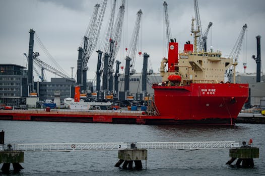A red cargo ship named 'Sun Rise' docked at an industrial port with cranes.
