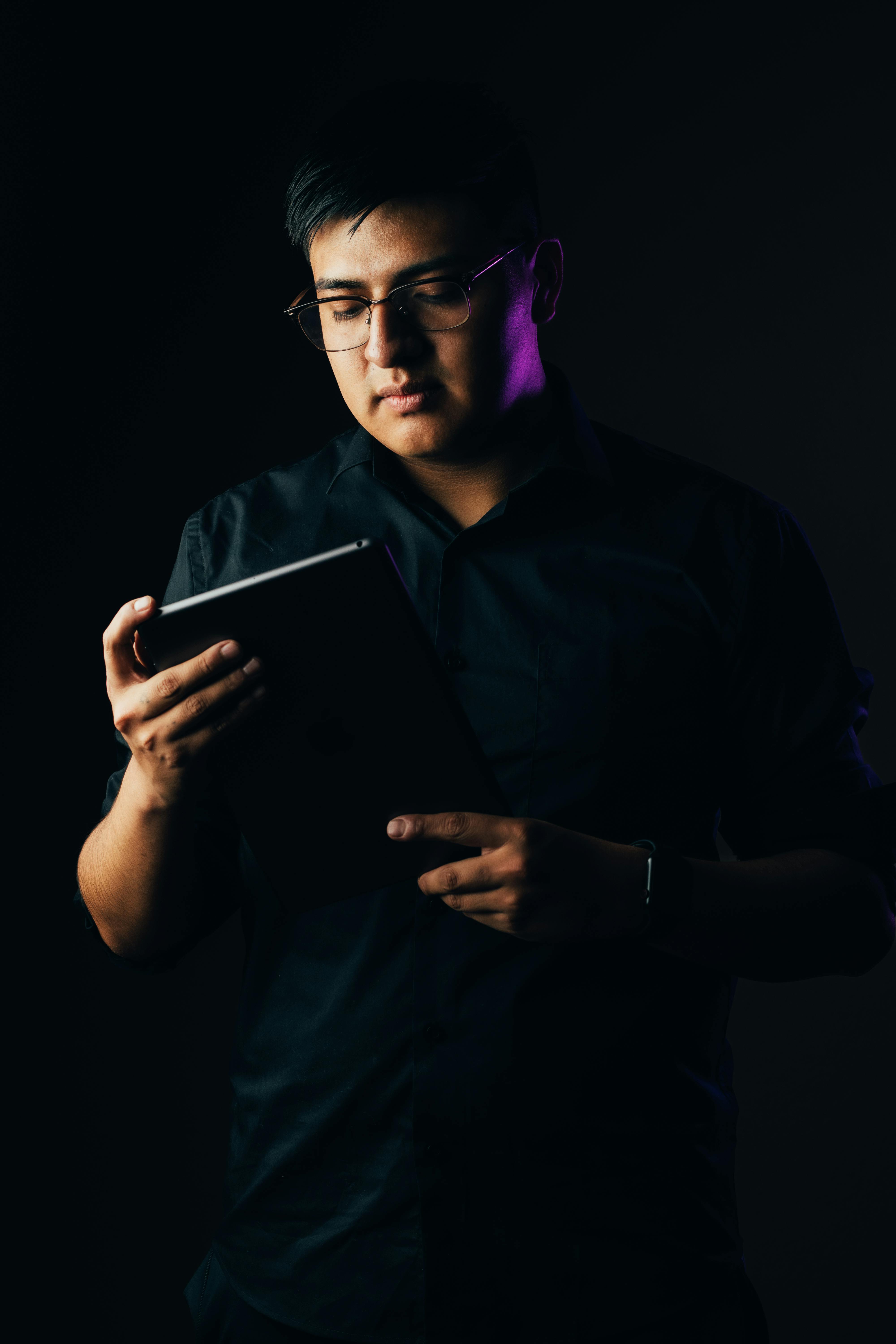 ฟรี ชายสวมแว่นตากำลังตั้งใจใช้แท็บเล็ตในสตูดิโอที่มีบรรยากาศหม่นหมอง คลังภาพถ่าย