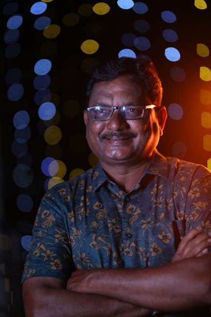 Smiling man in patterned shirt against colorful bokeh background, arms crossed.