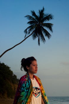 A woman wrapped in a vibrant shawl gazes at a sunset on a Venezuelan beach.