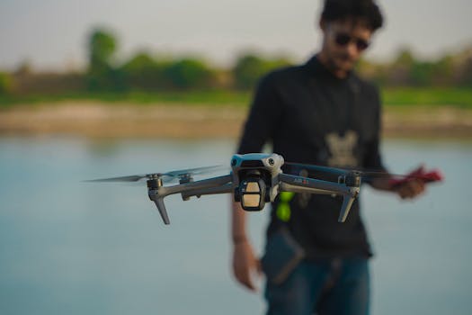 A drone in flight with a blurred background of a male operator by a river.