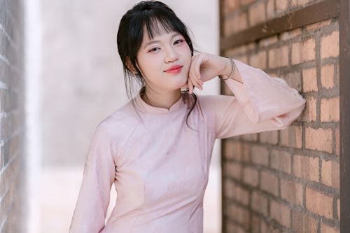 Free Portrait of a young woman in a pink traditional dress leaning against a brick wall, smiling warmly. Stock Photo