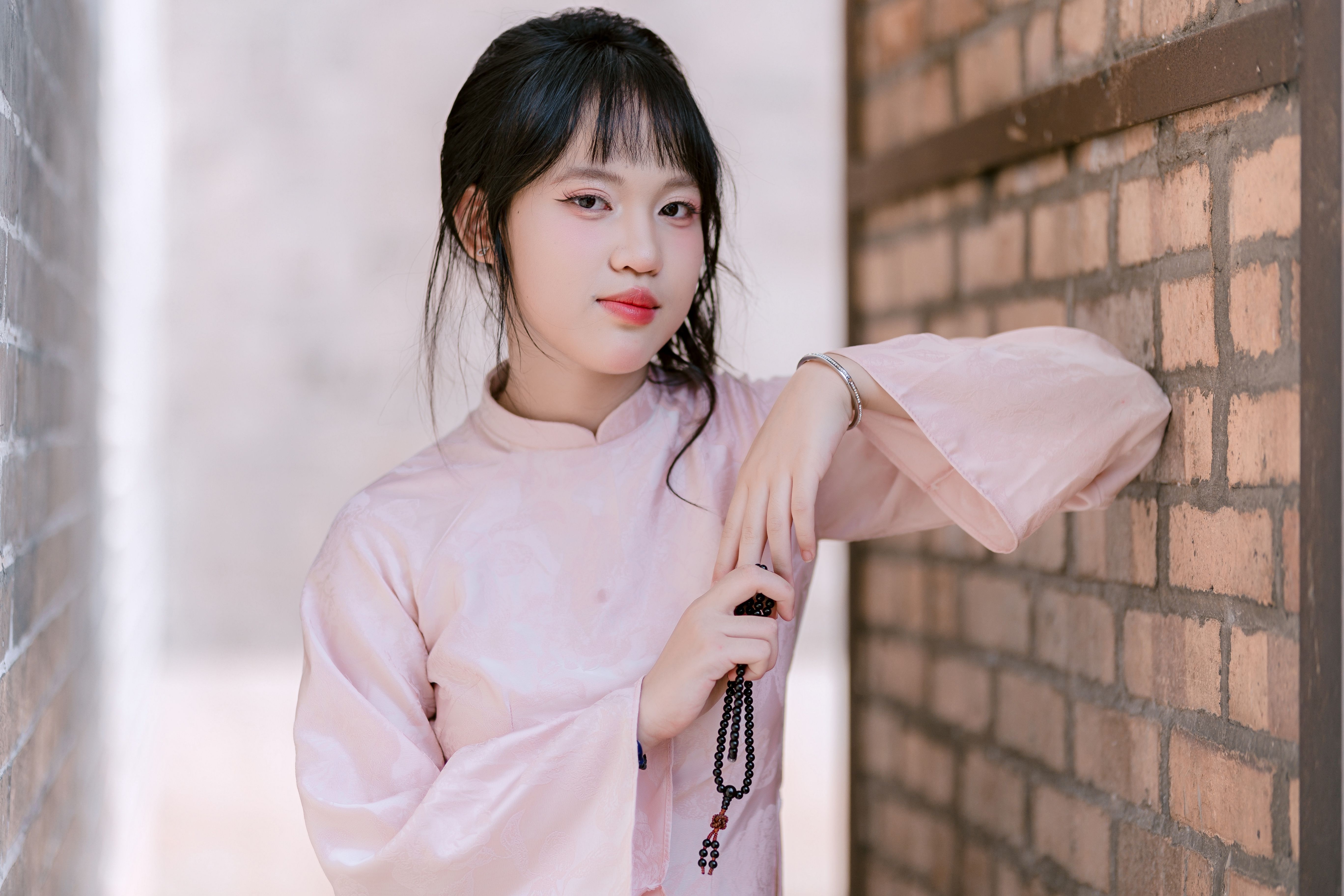 Portrait of a young woman in traditional pink dress holding a rosary, leaning against a brick wall.