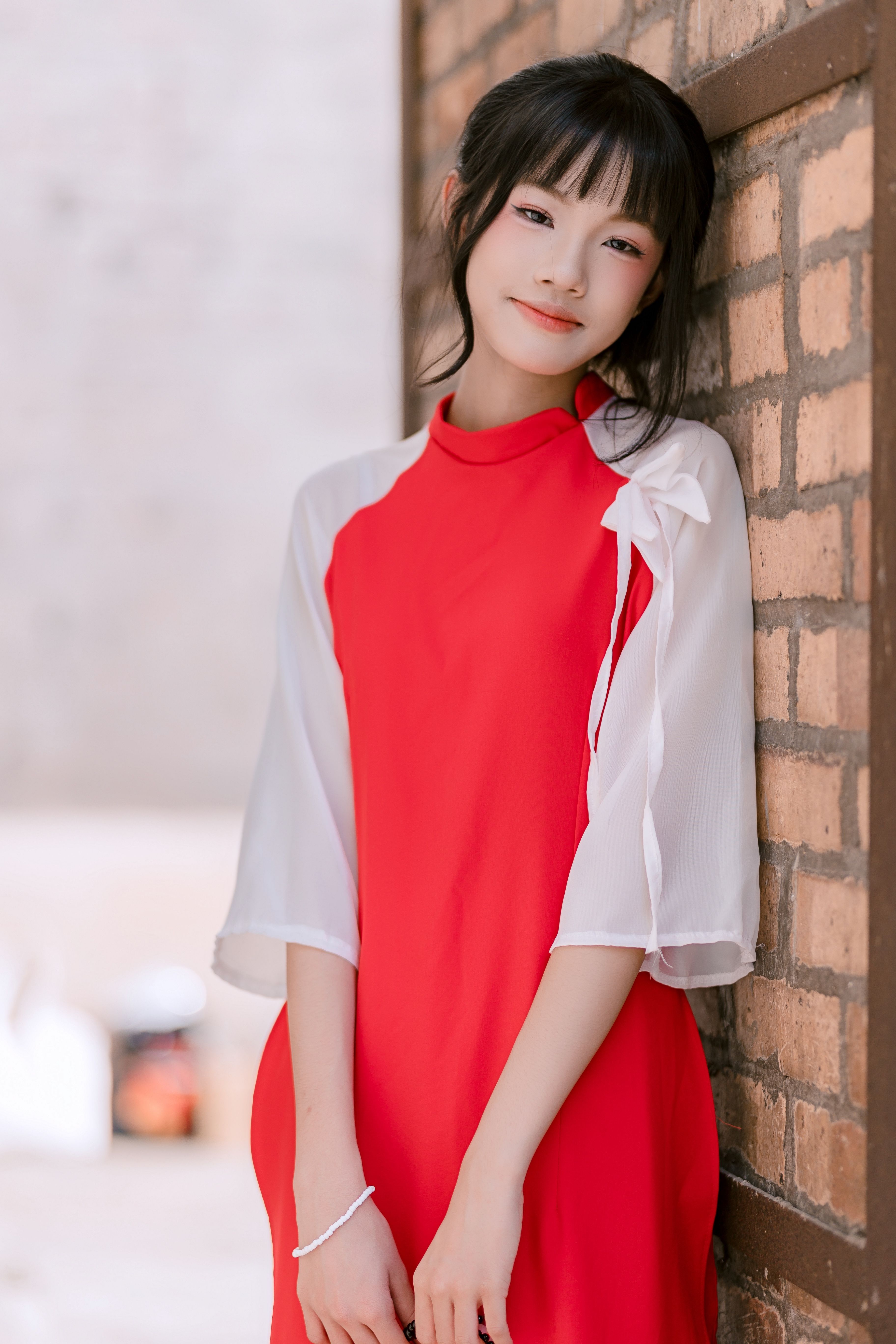 Young woman in a red dress leaning against a brick wall, exuding elegance and style.