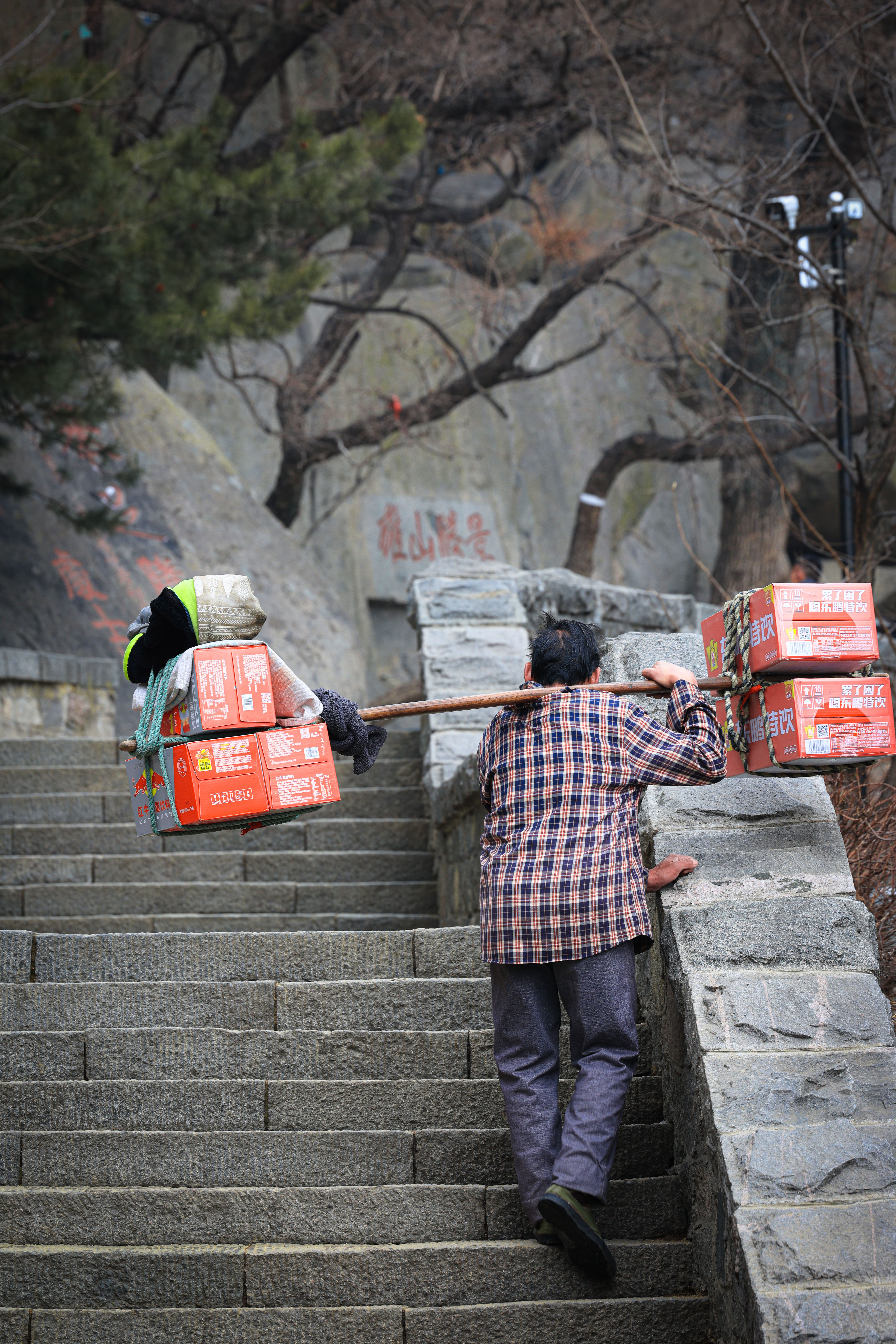 Elderly man carrying boxes on a pole up stone steps, showcasing resilience and tradition.