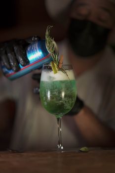 A bartender prepares a vibrant green cocktail with rosemary garnish in a dimly lit setting.