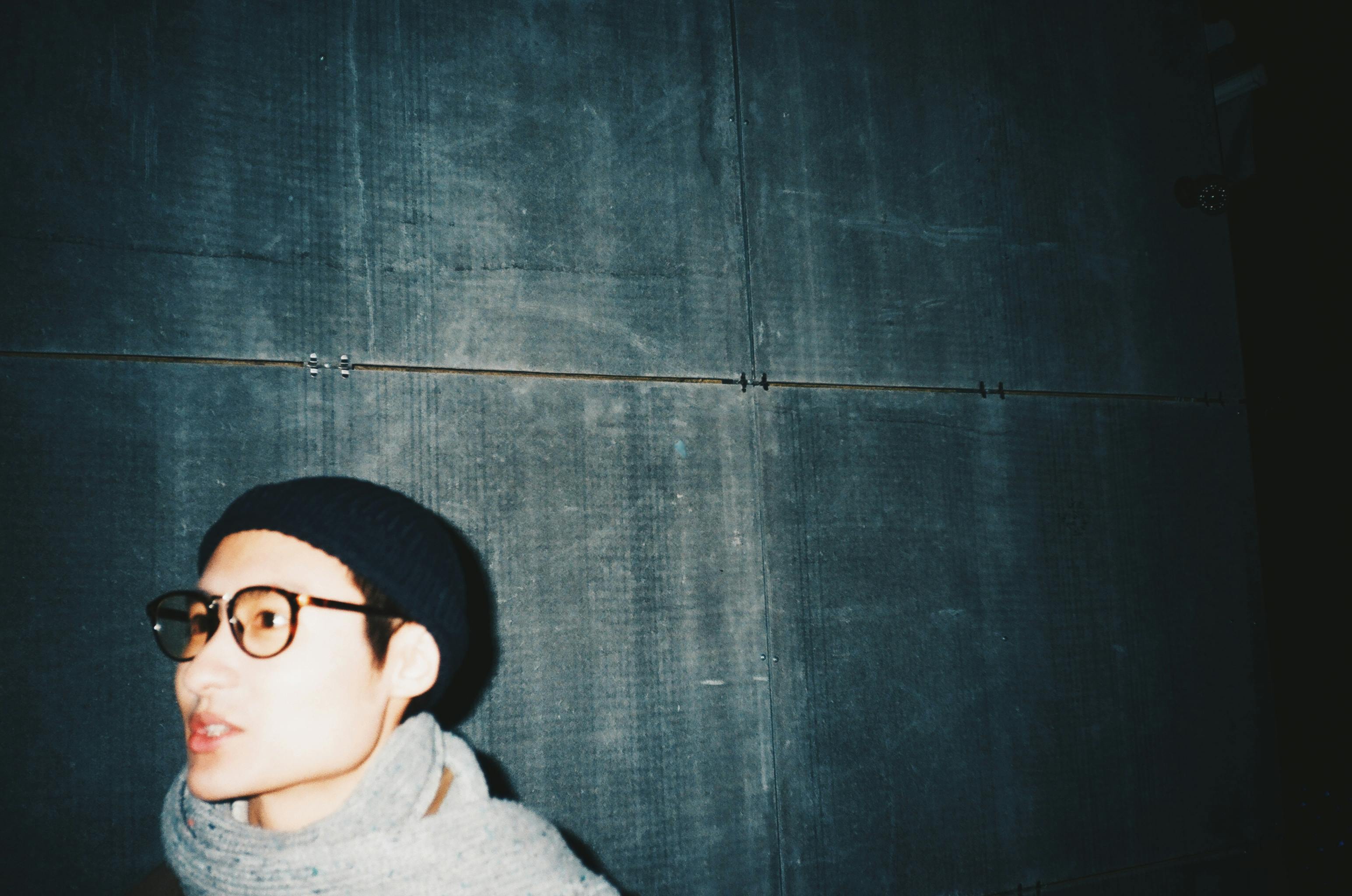 这是一幅艺术肖像，描绘了一位戴着眼镜和无檐帽的时尚成年人，背景是昏暗的城市环境