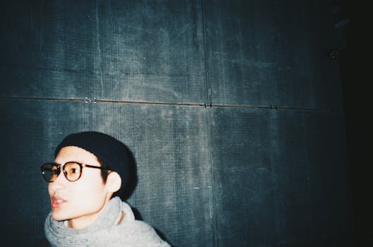 这是一幅艺术肖像，描绘了一位戴着眼镜和无檐帽的时尚成年人，背景是昏暗的城市环境