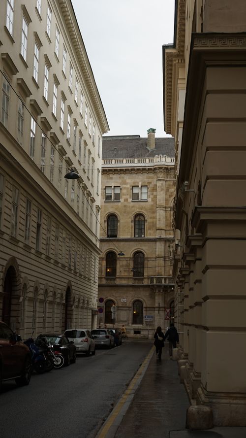 Free A narrow European street lined with historic buildings, featuring cars and people walking on a cloudy day. Stock Photo