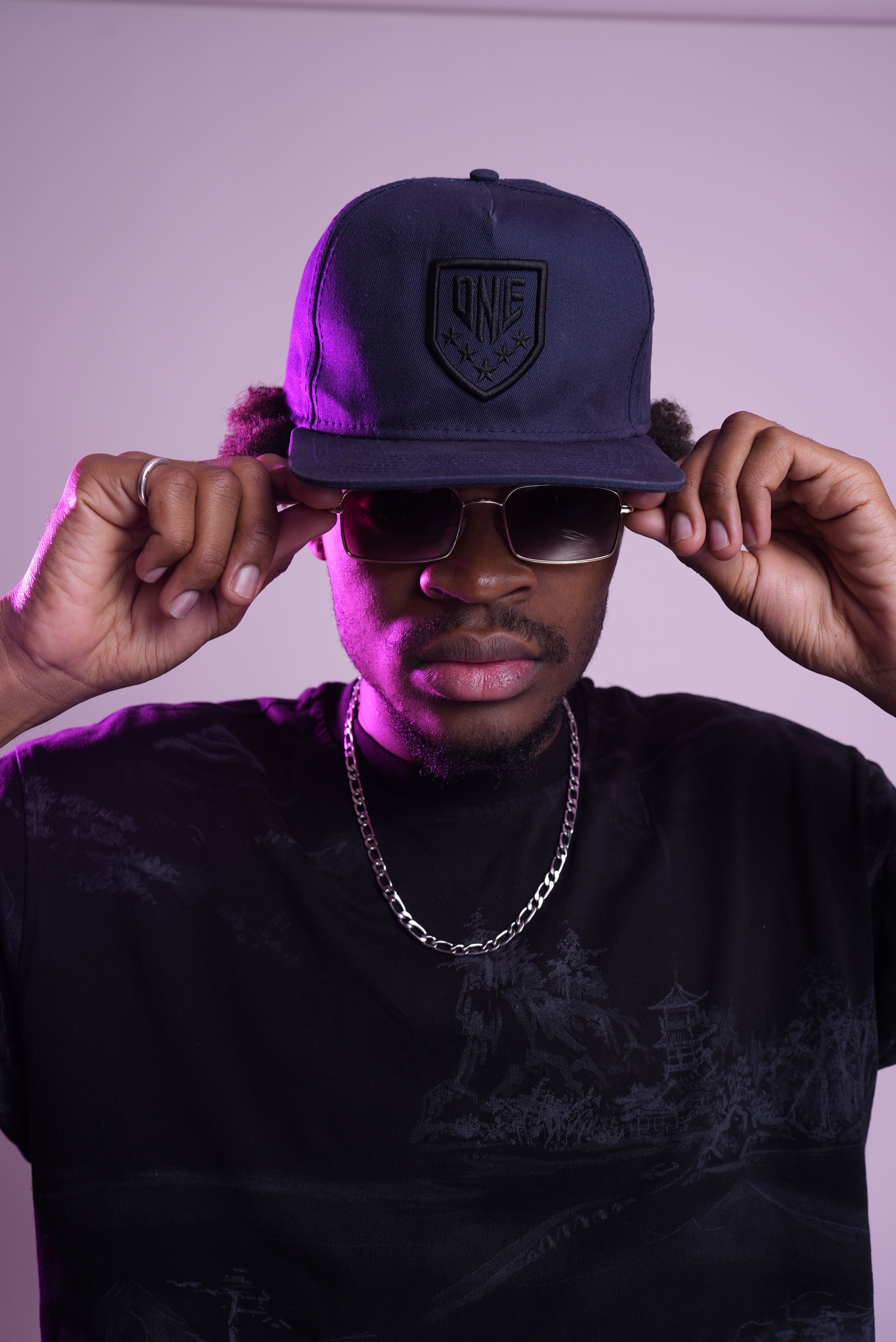 Young man posing with cap and sunglasses under purple lighting. Fashionable and confident style.