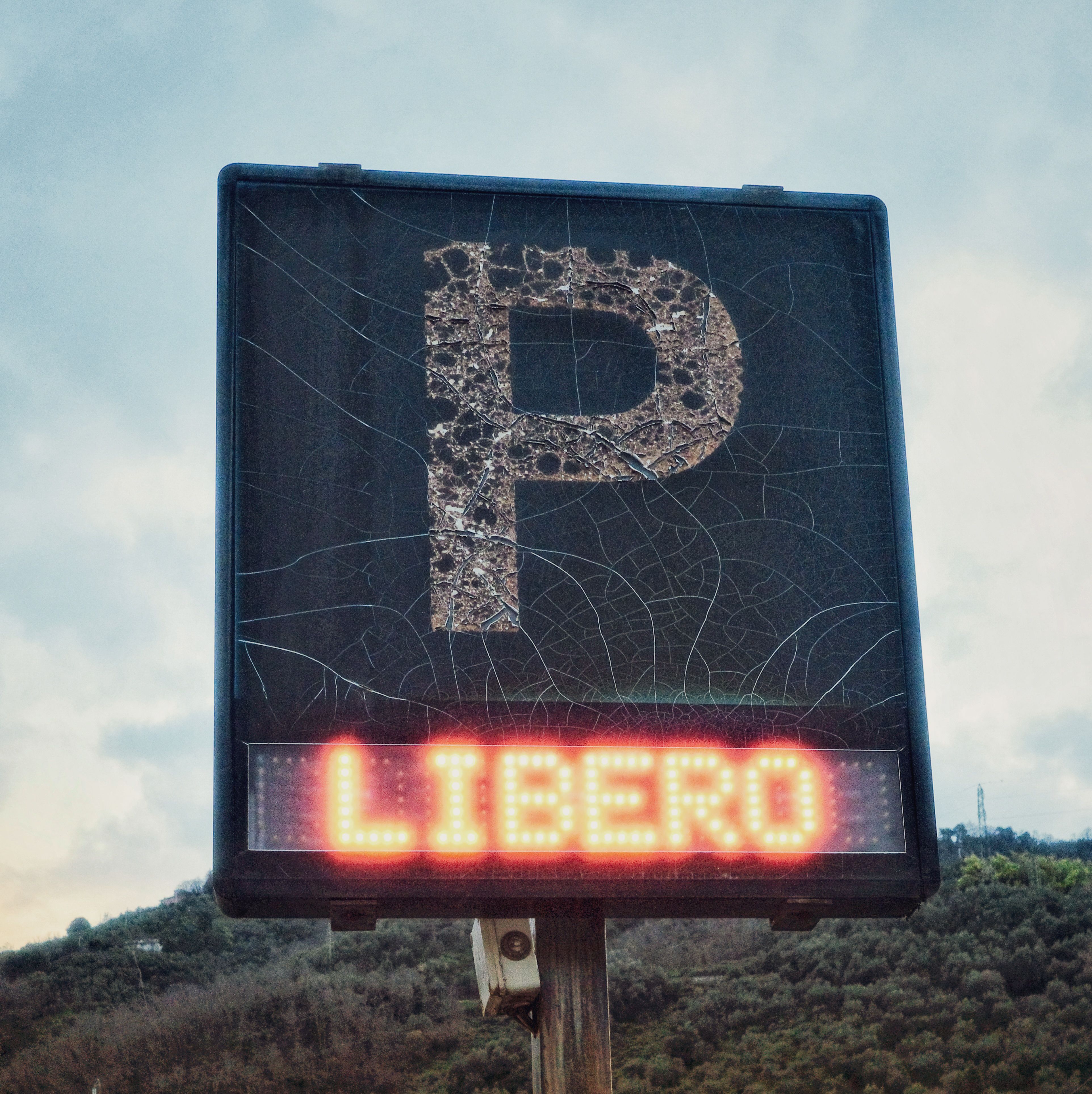 Old parking sign with crackled texture and 'LIBERO' lit in neon, outdoors in a natural setting.