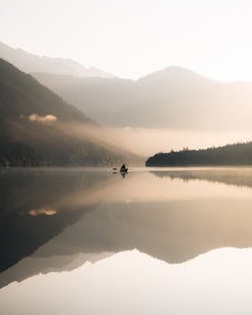 Tranquil scene of a lone canoe on a calm lake surrounded by misty mountains at sunrise.