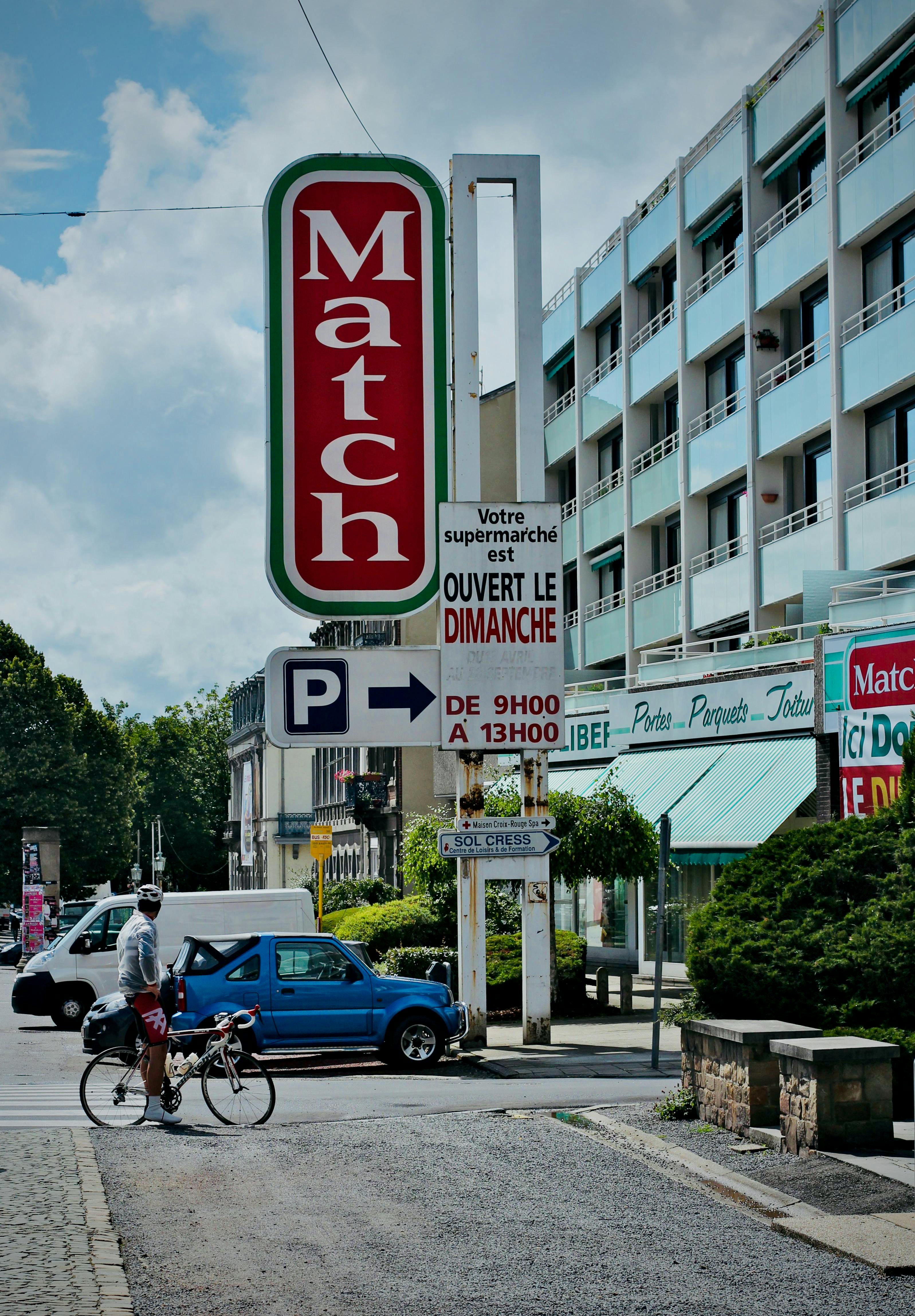 Free A city street view featuring a cyclist, car, and supermarket signs, creating an urban atmosphere. Stock Photo