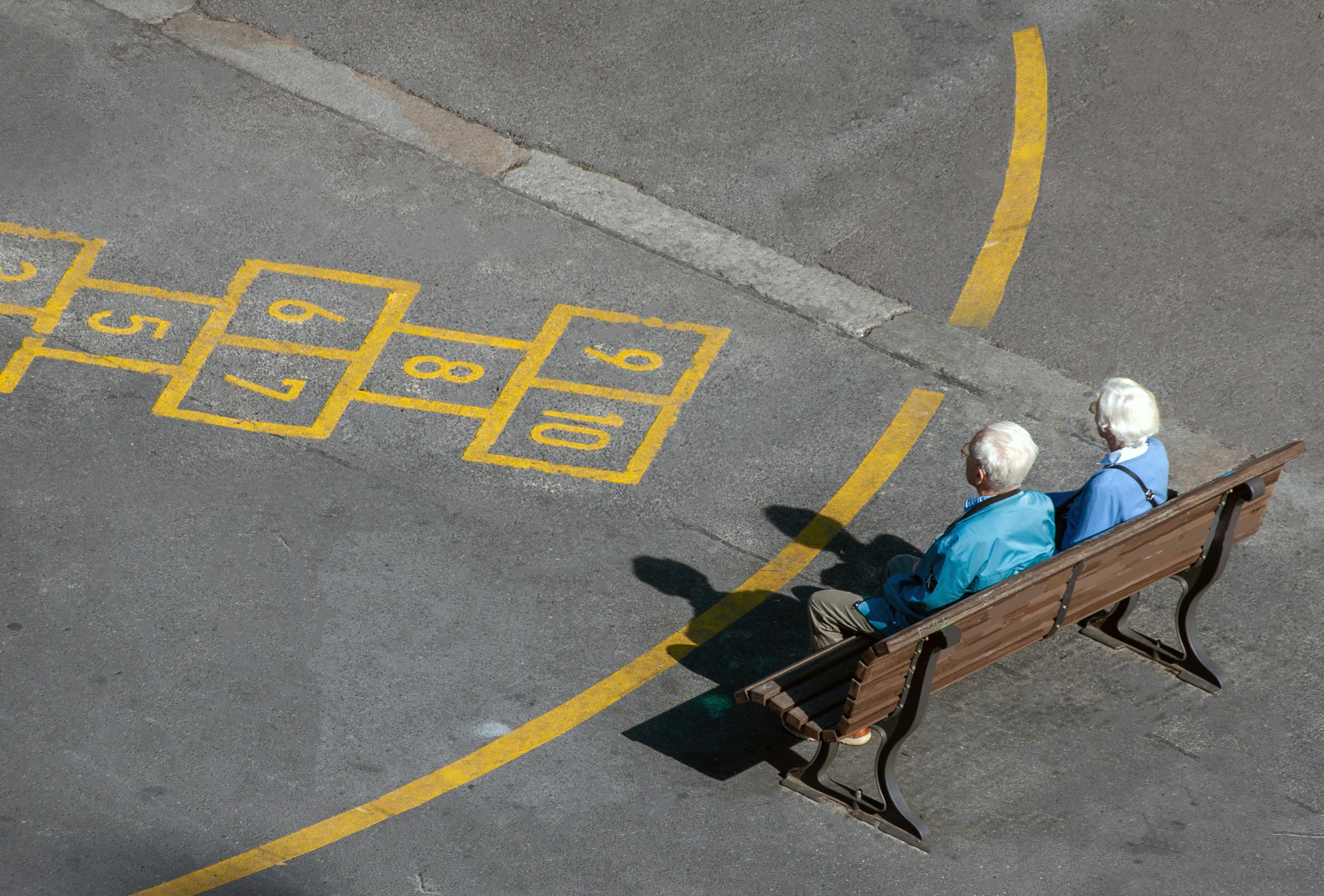 Gratis Et eldre par koser seg med en solrik dag på en parkbenk ved siden av en hopscotch-lek. Arkivbilde