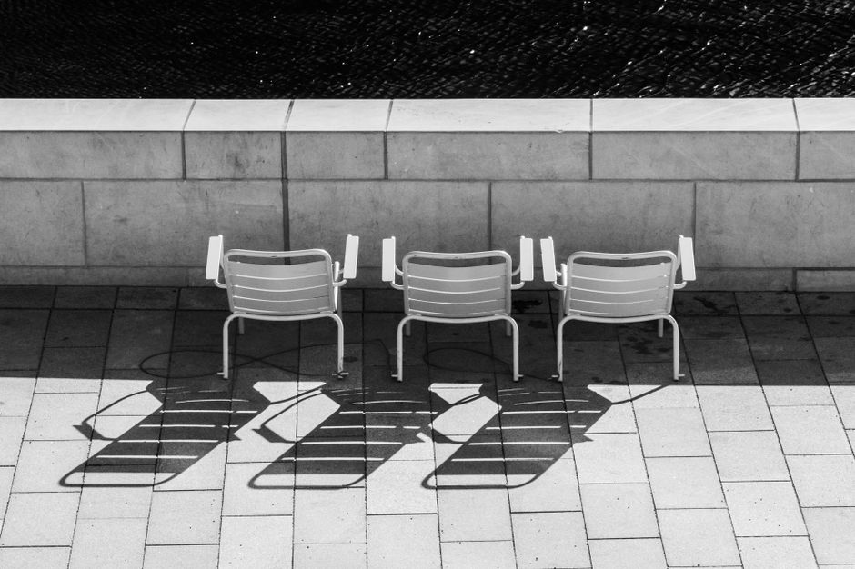 Black and white photo of three chairs casting long shadows on a sunlit pavement.