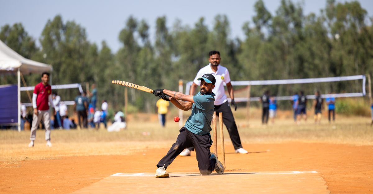 A cricket batter in motion swings a bat against a bright stadium backdrop under blazing floodlights.
