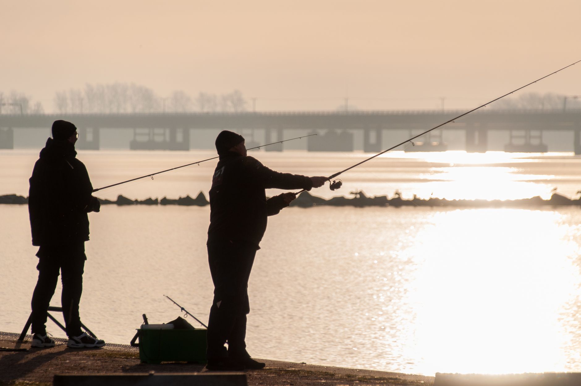 Two fishermen silhouetted against a sunrise as they cast their lines by the water.