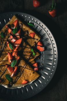 Appetizing crepes with strawberries, mint, and chocolate drizzle on a dark plate.