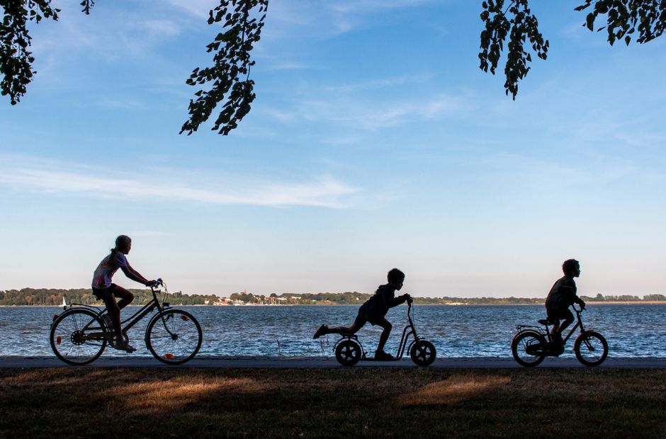 Silhouettes of children playing on bikes and scooter by a serene lakeside during daylight.