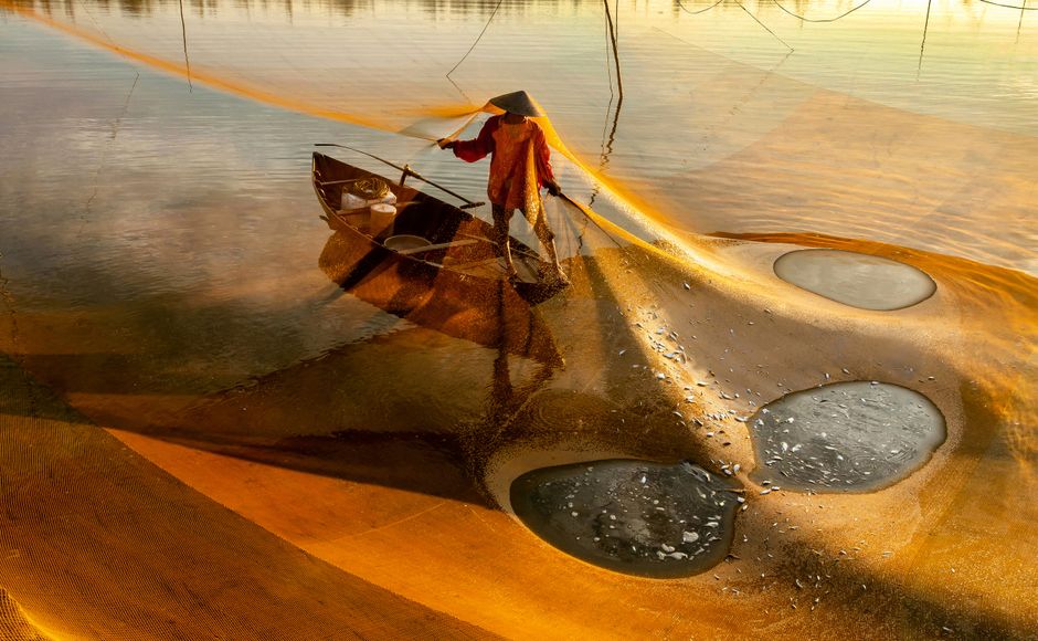 A Vietnamese fisherman casting a net from a boat on a calm river, captured in warm evening light.