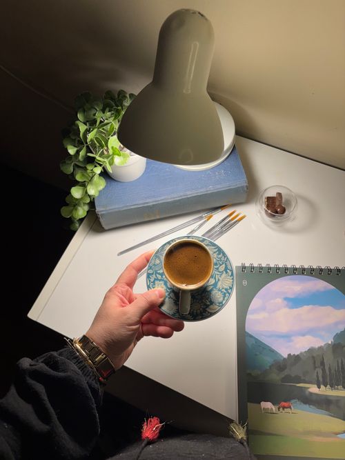 Free A hand holds a coffee cup on a well-lit desk with sketchbook and lamp. Stock Photo