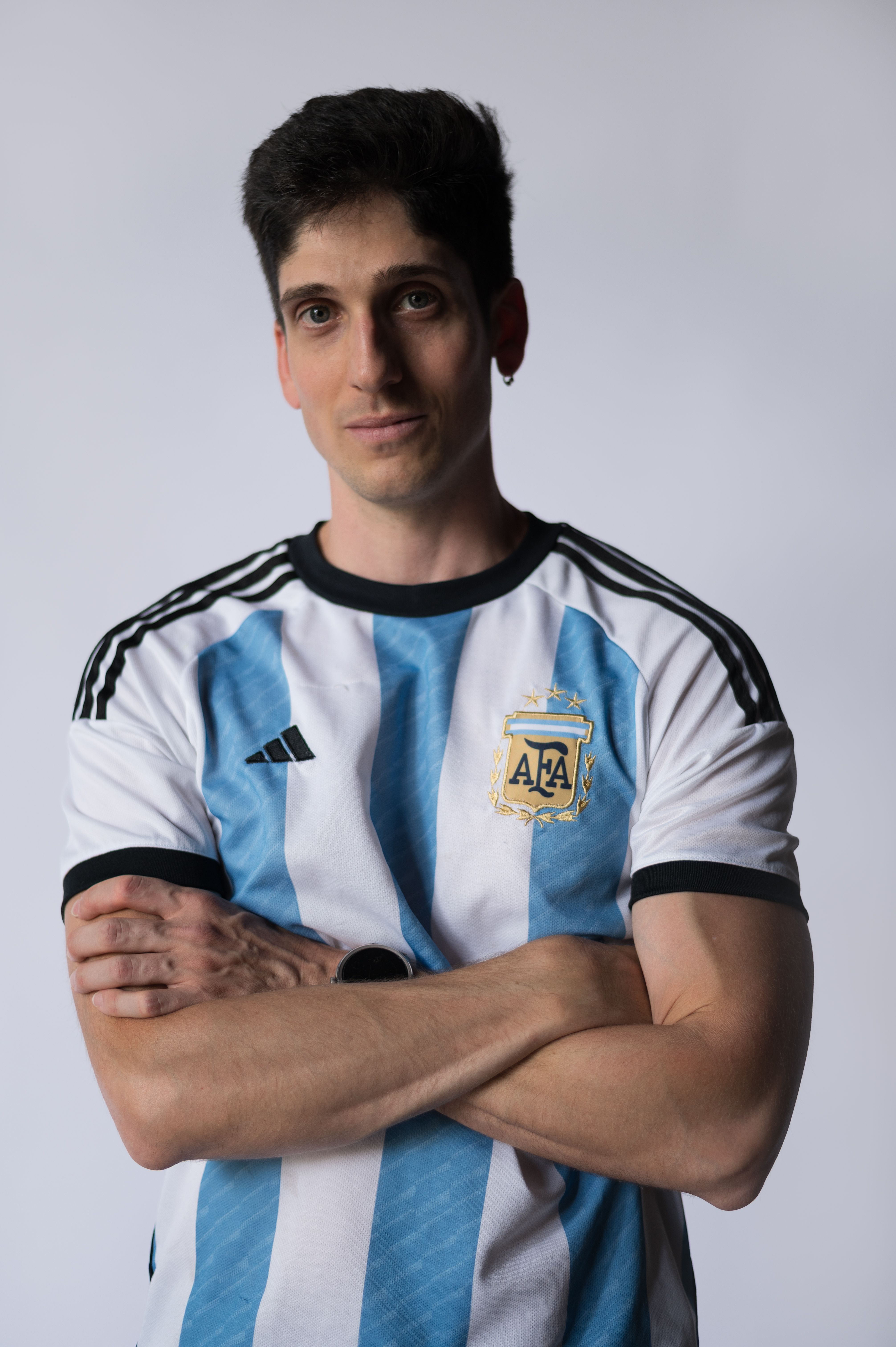 Portrait of a man in an Argentina soccer jersey, arms crossed confidently indoors.