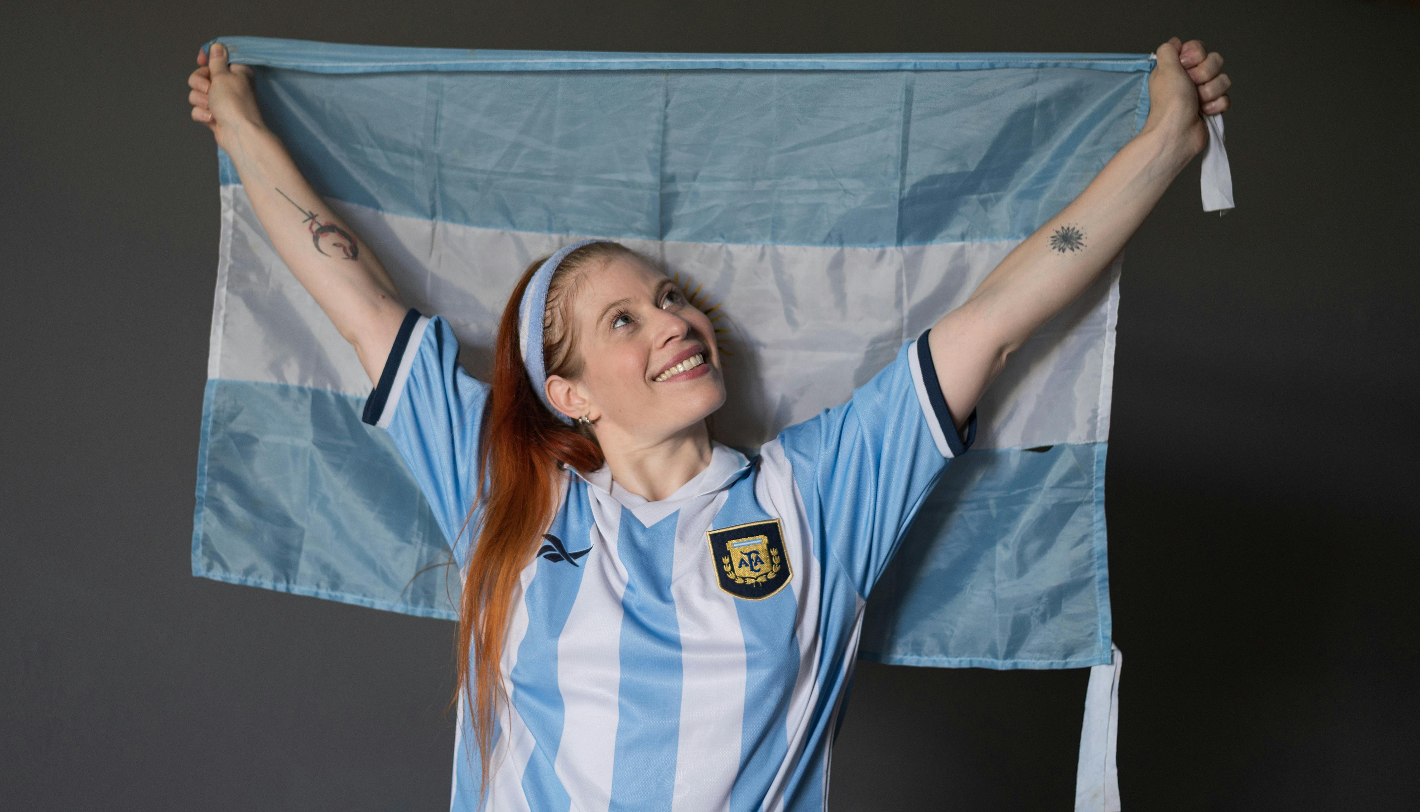 Joyful woman holding Argentina flag wearing national jersey indoors.