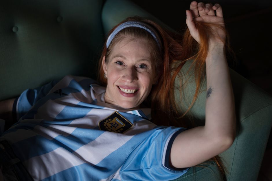 Cheerful woman in an Argentina soccer jersey lying on a couch, enjoying a relaxed moment indoors.