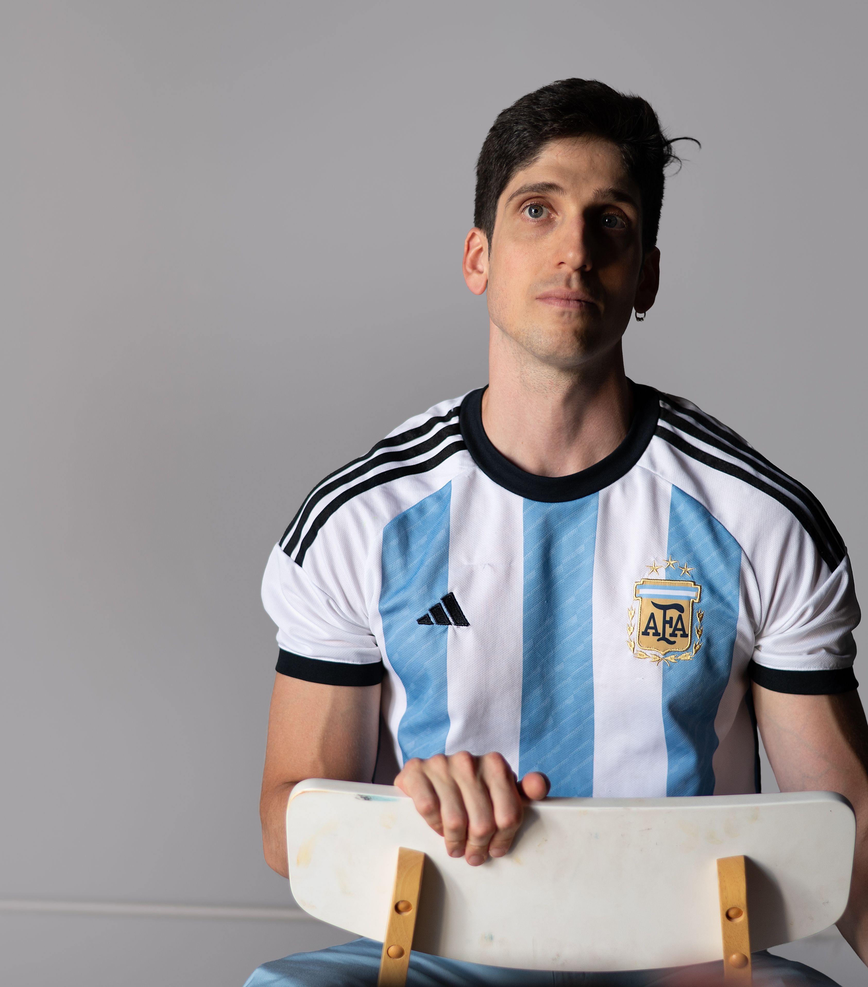 Portrait of a young man wearing an Argentina football jersey seated indoors on a minimal background.