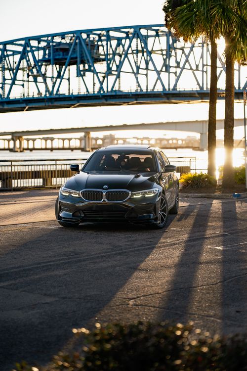 Free A sleek luxury car parked near a river at sunset with a iconic blue bridge in the background. Stock Photo